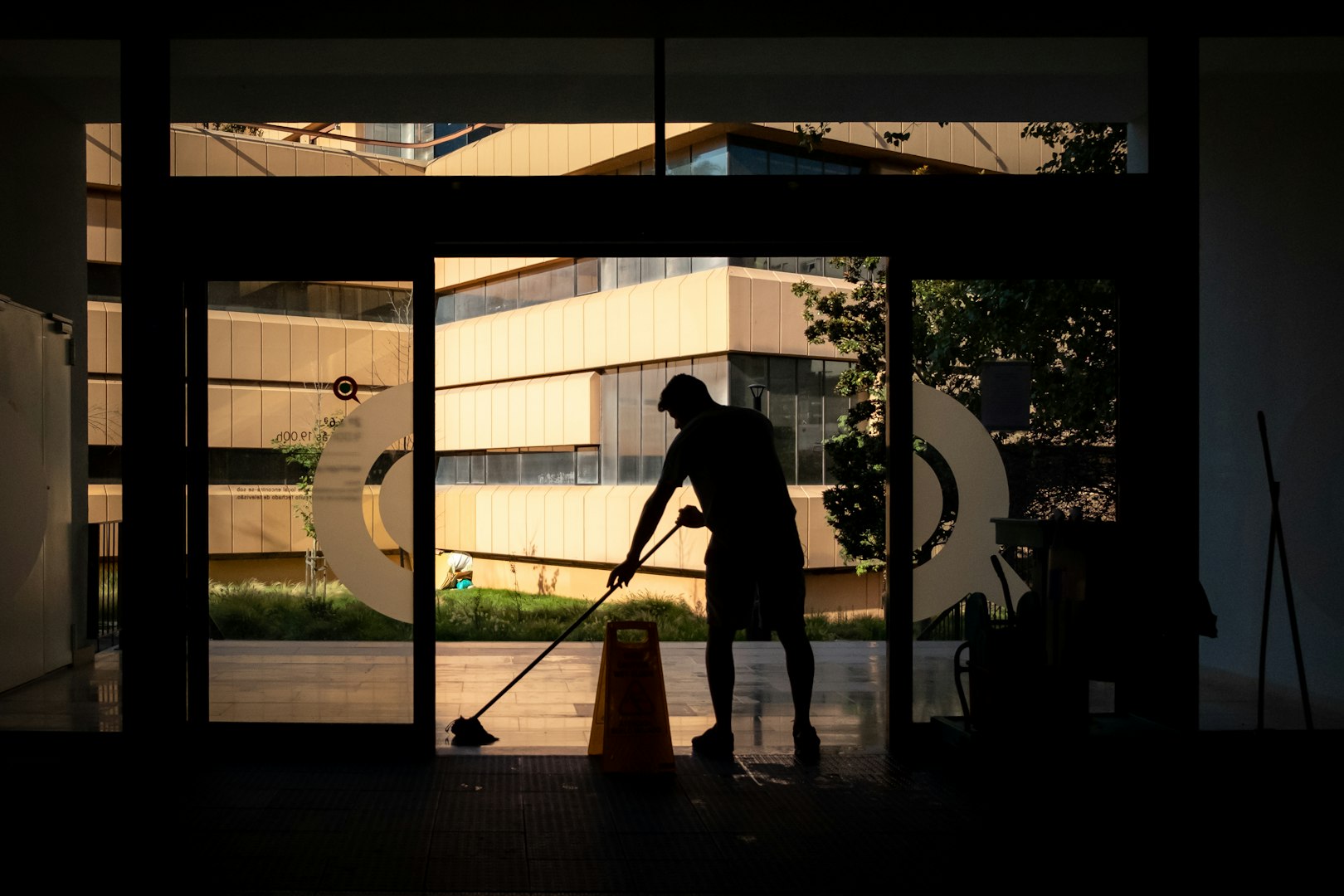 Aerial view of a cleaning crew in a modern office space, with two individuals using mops and a vacuum cleaner, and one person cleaning stairs. All are dressed in blue overalls and wearing yellow gloves. The environment features plants and contemporary furniture.
